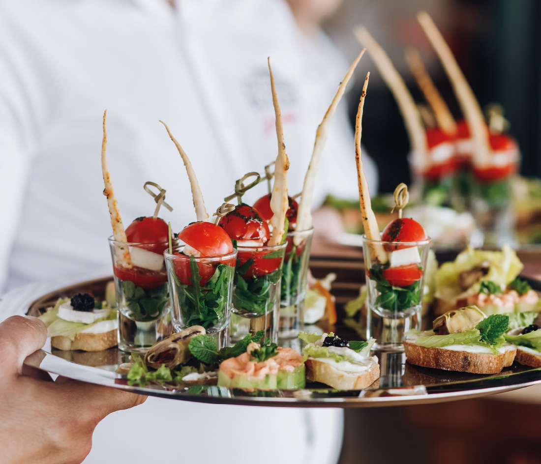 Ein Kellner hält ein Tablett mit Vorspeisen: Gläser gefüllt mit Kirschtomaten, Gemüse und Sticks, daneben belegte Sandwiches mit verschiedenen Zutaten wie Lachs, Kaviar und Kräutern. Der Hintergrund ist unscharf.