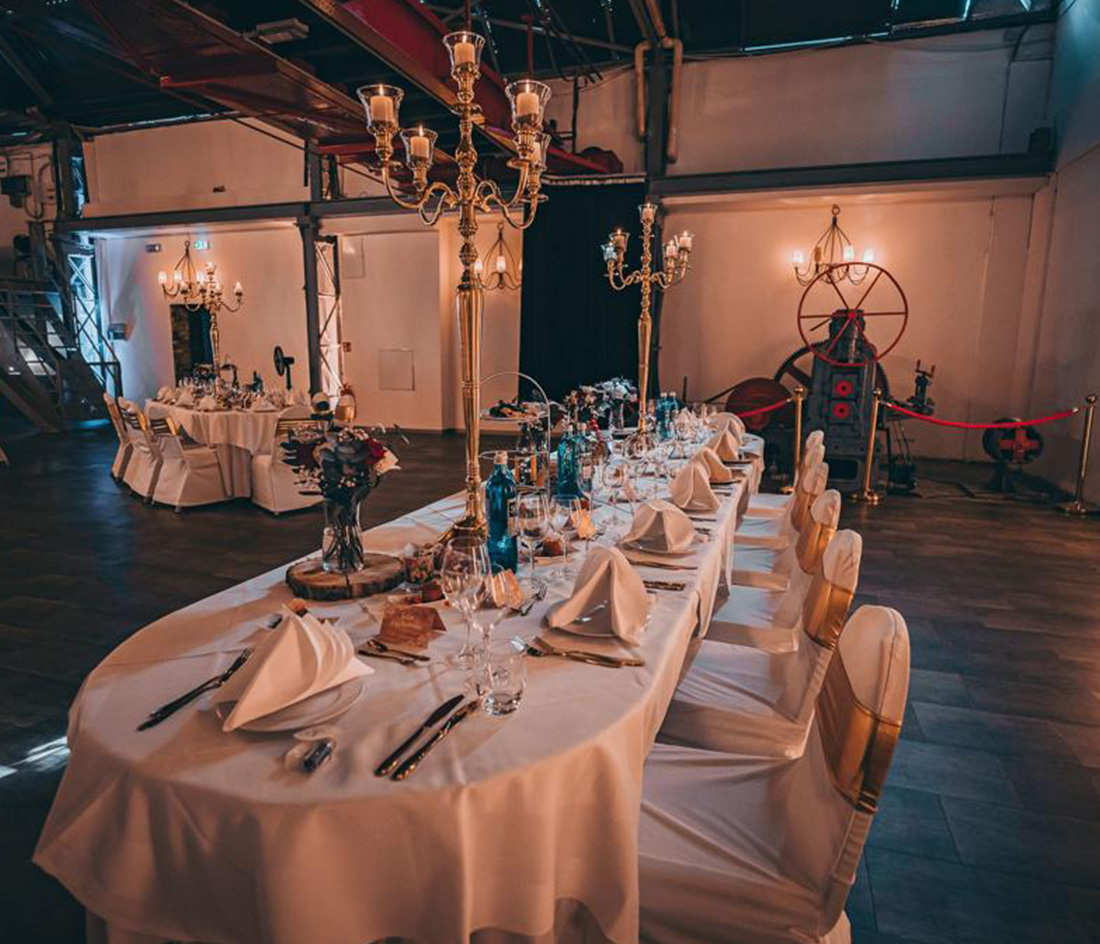 Ein wunderschön dekorierter Bankettsaal mit elegant gedeckten Tischen. Hohe Kandelaber dienen als Tischdekoration, dazu weiße Tischdecken und gefaltete Servietten. Stimmungsvolle Beleuchtung sorgt für eine warme Atmosphäre, und dekorative Elemente schmücken den großzügigen Saal.