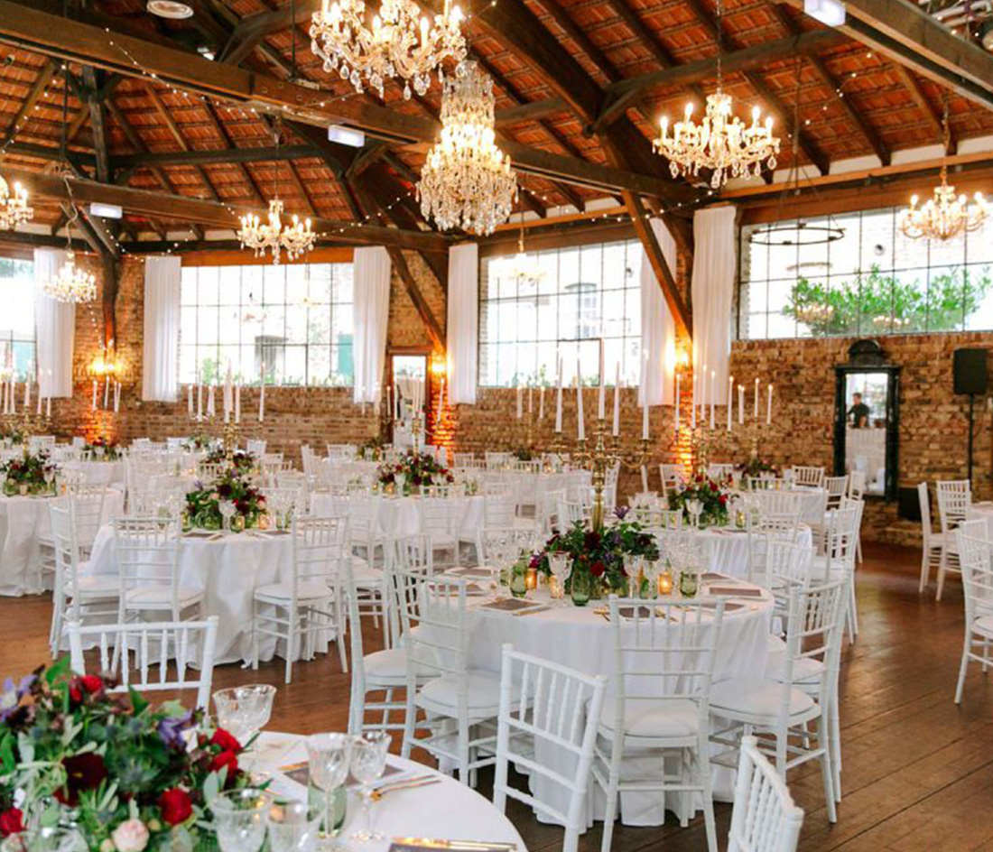 Geräumiger Veranstaltungssaal mit Holzbalken und Backsteinwänden, ausgestattet mit runden Tischen mit weißen Stühlen und Tischdecken. Jeder Tisch ist mit Blumenschmuck geschmückt. Kronleuchter und hohe Fenster schaffen eine helle, elegante Atmosphäre.