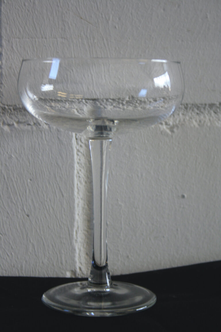 Das Champagnerkelch ist ein klares Cocktailglas mit breiter, flacher Schale und schlankem Stiel, das vor einer strukturierten grauen Wand auf einer schwarzen Oberfläche steht.