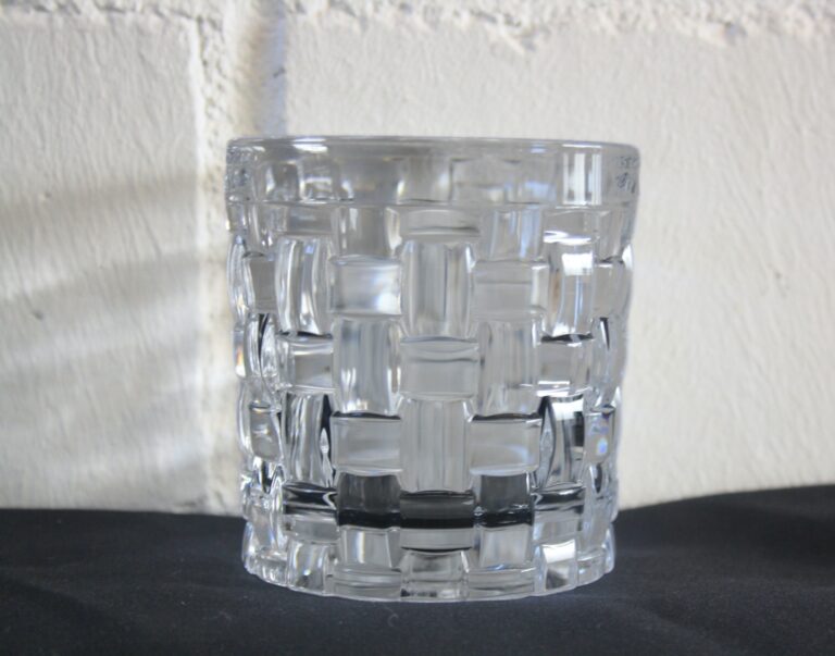 Das Cocktailglas Kristall mit seinem strukturierten Schachbrettmuster steht auf einer schwarzen Oberfläche vor einer strukturierten weißen Wand.