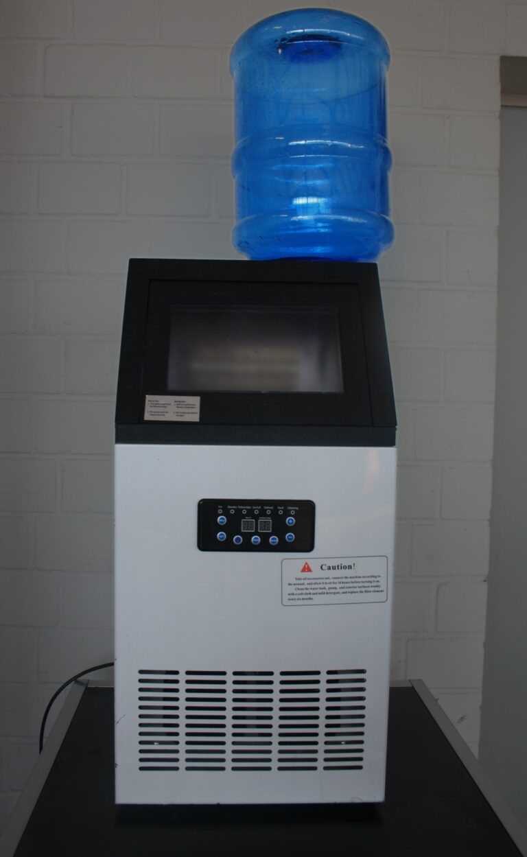 Eine weiße Eiswürfelmaschine (72 kg/24 h) mit einem großen blauen Wasserkrug obenauf verfügt über ein digitales Bedienfeld auf der Vorderseite und steht vor einer weißen Ziegelwand.
