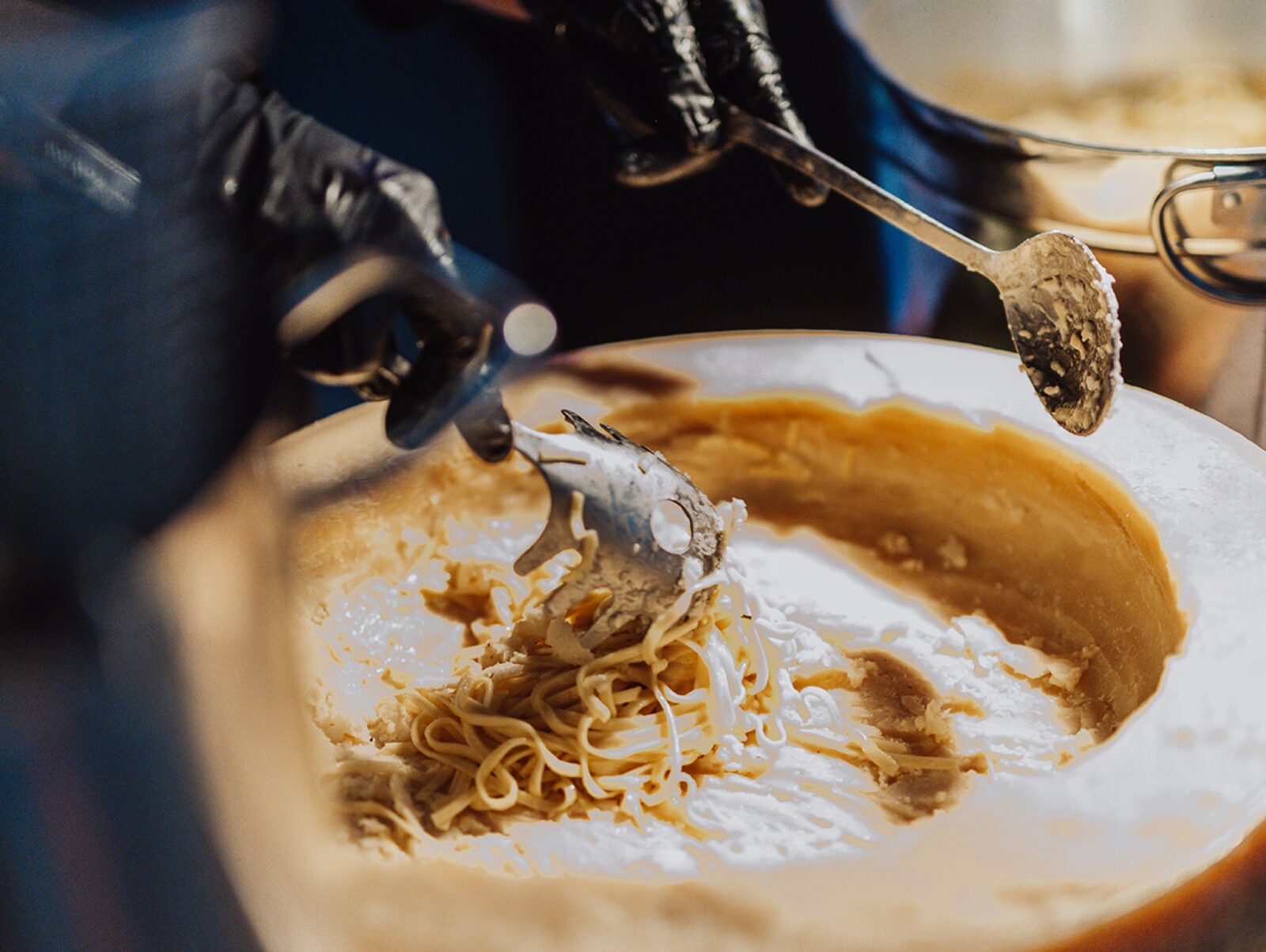 Eine Person mit schwarzen Handschuhen rührt mit Gabel und Löffel Spaghetti in einem großen Käselaib um. Die Spaghetti scheinen mit Soße überzogen zu sein. Im Hintergrund ist ein Metalltopf teilweise sichtbar.