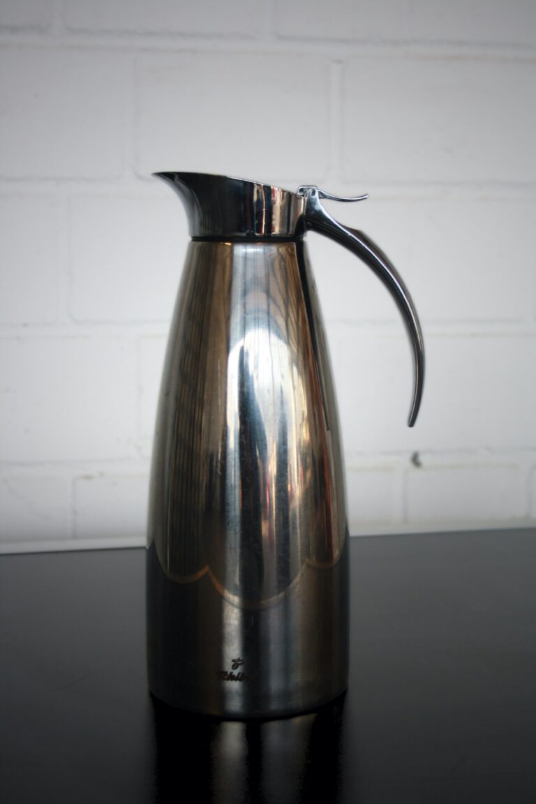 Die Kaffeekanne Silber, eine elegante, reflektierende Metallkanne mit gebogenem Griff und Ausguss, steht auf einer dunklen Oberfläche vor dem Hintergrund einer weißen Ziegelwand.