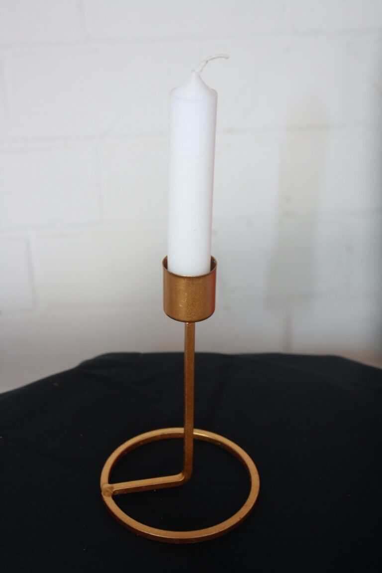 Eine weiße Kerze ruht im Kerzenständer Lunavia, einem schlichten, modernen Metallhalter mit Goldfinish. Er verfügt über einen runden Sockel und Stiel und steht auf einer dunklen Stoffoberfläche vor einem hellen Wandhintergrund.