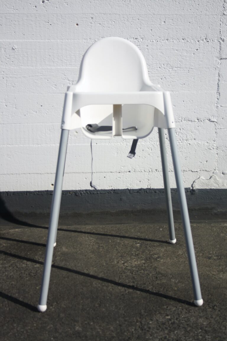 Der Kinderstuhl, ein weißer Hochstuhl mit minimalistischem Design und grauen Metallbeinen, steht vor einer hellgrauen Betonwand. Sein Schatten fällt teilweise auf den dunkelgrauen Boden.