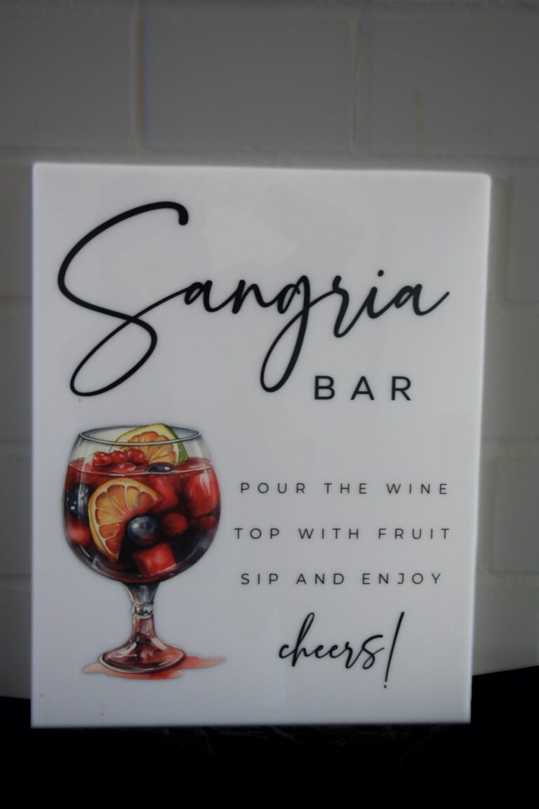 Das Sangria-Schild zeigt ein Glas mit Sangria, Fruchtscheiben und Beeren. Der Text lautet: „Wein einschenken, mit Früchten garnieren, nippen und genießen. Prost!“ vor einer hellen Backsteinwand.