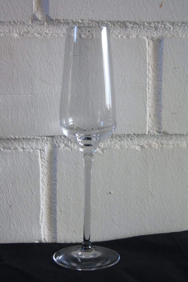 Ein hohes, leeres Sektglas mit schlankem Stiel steht vor dem Hintergrund einer weißen Ziegelwand, klar und auf einer schwarzen Oberfläche ruhend.