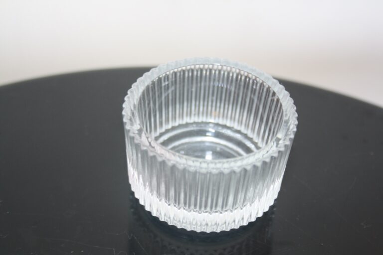 Das Teelichtglas Elara, eine klare, gerippte Glasschale mit vertikalen Rillen, die ihre zylindrische Form betonen, steht auf einer glänzend schwarzen Oberfläche. Das Licht wirft einen subtilen Reflex darauf.