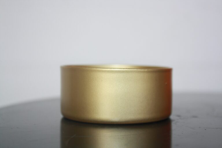Das Teelicht Gold Mirabella, ein kleiner, glatter, metallisch goldener Zylinder, ruht auf einer reflektierenden dunklen Oberfläche. Seine Schlichtheit und Eleganz werden durch den schlichten, zartweißen Hintergrund hervorgehoben.