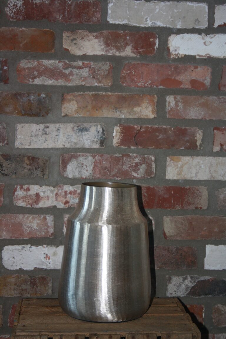 Die Vase Gold Lumis mit ihrem schlichten Design steht auf einer Holzkiste. Dahinter befindet sich eine Ziegelwand in verschiedenen Rot-, Braun- und Grautönen.